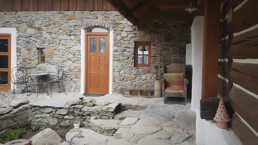 Kamenná stěna domku, dřevěné dveře, kovový stolek a křeslo na kamenné dlažbě